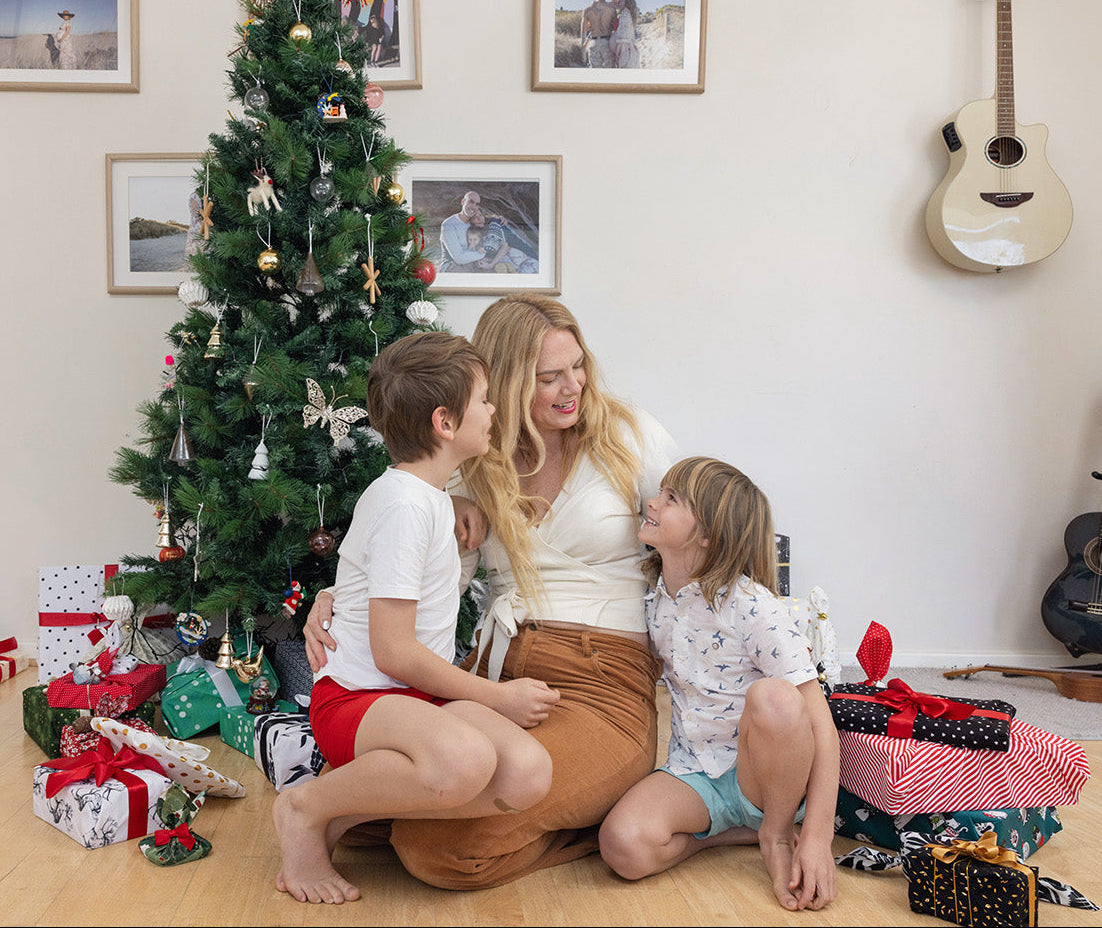 Woman and two children sitting on the floor in front of a Christmas tree with presents. Tis the Season Xmas Prints. Festive Kit.